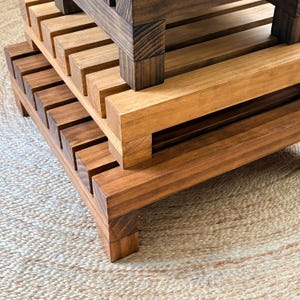 May include: A tiered wooden stand constructed from horizontal slats of varying brown wood tones. The structure features a dark espresso-colored top tier and a lighter, medium-brown lower tier, both supported by sturdy, block-style wooden legs.