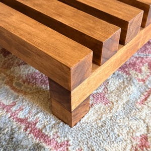 May include: A close-up of a wooden plant stand with a warm brown finish. The stand features a slatted top and sturdy legs, showcasing the natural wood grain. The design is simple and modern, suitable for displaying plants or decorative items.