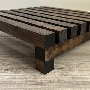 May include: A dark stained wooden plant stand with a slatted top and square legs. The stand is made of solid wood and has a rich, brown color. The design is simple and modern, suitable for displaying plants or other decorative items.