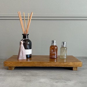 May include: A wooden tray with a diffuser bottle, a tassel, and two small bottles of body wash. The diffuser bottle is brown glass with a clear glass lid and wooden sticks. The tassel is pink and white. The body wash bottles are clear glass with silver lids and labels that say "Molton Brown".