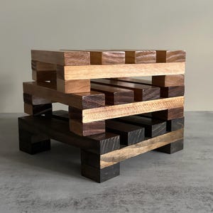 May include: A stack of four wooden coasters, each with a different wood grain pattern. The top two coasters are light brown, the middle coaster is a darker brown, and the bottom coaster is a dark brown with a black stain.