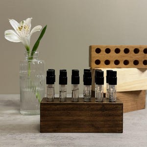 May include: A display of small glass vials with black caps, arranged in a dark wooden holder. A clear glass vase with a white flower and a wooden block with holes are in the background. The vials appear to contain liquid, possibly perfume samples.