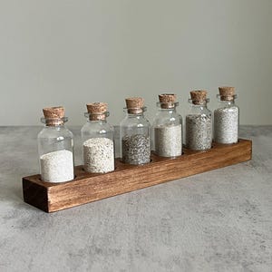 May include: A wooden spice rack with six small glass bottles, each filled with a different type of spice. The bottles have cork stoppers and are arranged in a row. The rack is stained a dark brown color.