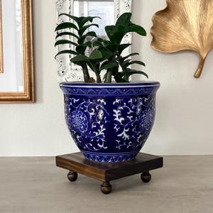 May include: A deep blue ceramic planter featuring a white floral vine pattern and geometric border designs. The round pot sits on a square dark brown wooden stand with four small ball-shaped feet, containing a green ZZ plant with thick stems.