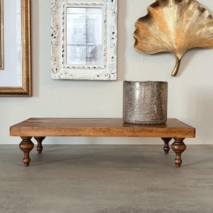 May include: A rectangular wooden riser with a medium brown finish, supported by four turned spindle legs. A cylindrical, textured metallic silver vase with a weathered, antique-style finish sits on the right side of the wooden platform surface.
