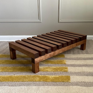 May include: A wooden footstool with a slatted top and sturdy legs. The footrest is made of dark brown wood, with a natural finish. The footstool measures approximately 24 inches long, 12 inches wide, and 8 inches high.
