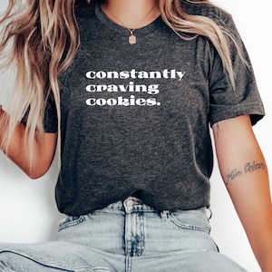 Könnte beinhalten: Ein dunkelgraues T-Shirt mit dem Aufdruck "constantly craving cookies." in Weiß.