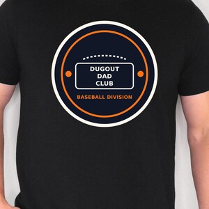 May include: Black t-shirt with a circular graphic. The graphic features a navy blue circle with an orange outline and accents. Inside the circle is a white rectangle with the words "DUGOUT DAD CLUB" and "BASEBALL DIVISION".