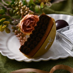 May include: A yellow and brown makeup brush with the name "Naomi" printed on the side, resting on a white plate. A clear perfume bottle and a dark chocolate candy are also on the plate, with a floral arrangement in the background.