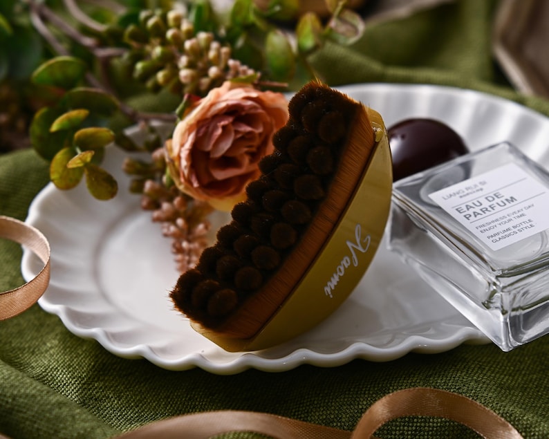 May include: A yellow and brown makeup brush with the name "Naomi" printed on the side, resting on a white plate. A clear perfume bottle and a dark chocolate candy are also on the plate, with a floral arrangement in the background.