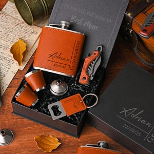 May include: A personalized groomsmen gift set featuring a brown leather-wrapped flask, shot glasses, a knife, a bottle opener, and a keychain. The set includes a gift box with the name "Adrian" and the date "22/10/25" engraved on the flask and box.