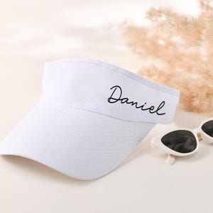 Peut inclure: Une visière blanche avec un nom brodé en noir "Daniel" sur le devant. La visière a un design en maille et est faite d'un matériau léger.