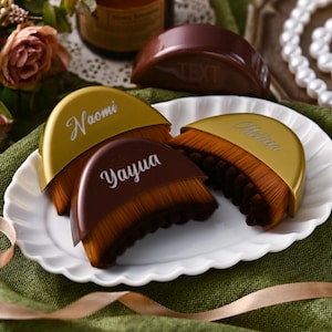 May include: A collection of makeup brushes with brown bristles and gold or brown handles, arranged on a white plate. The handles are inscribed with names in white script. A ribbon and flowers are in the background.
