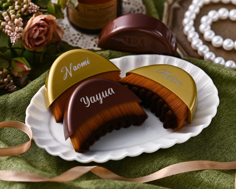 May include: A collection of makeup brushes with brown bristles and gold or brown handles, arranged on a white plate. The handles are inscribed with names in white script. A ribbon and flowers are in the background.