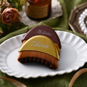 May include: Two personalized makeup brushes with brown bristles and curved handles. One handle is gold with the name "Shiran" and the other is brown with the name "Melissa". The brushes are on a white scalloped plate.