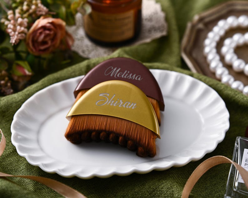 May include: Two personalized makeup brushes with brown bristles and curved handles. One handle is gold with the name "Shiran" and the other is brown with the name "Melissa". The brushes are on a white scalloped plate.