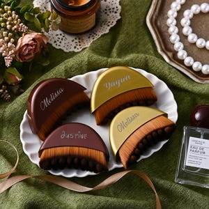 May include: Four makeup brushes with brown and gold handles, each with a name printed on them. The brushes are arranged on a white plate, with a perfume bottle and a string of pearls in the background. The brushes have a two-tone bristle design.
