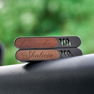 Könnte beinhalten: Zwei hölzerne Auto-Lufterfrischer mit Metallclips. Der obere Lufterfrischer ist braun und sagt "Nicole". Der untere Lufterfrischer ist hellbraun und sagt "Shalicia".