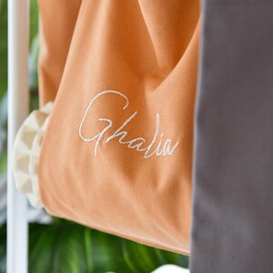 May include: Close-up of an orange canvas bag with the name "Ghalia" embroidered in white script. The bag has a ruffled, off-white trim and is partially visible, with a gray fabric in the background. The bag is hanging on a white rack.