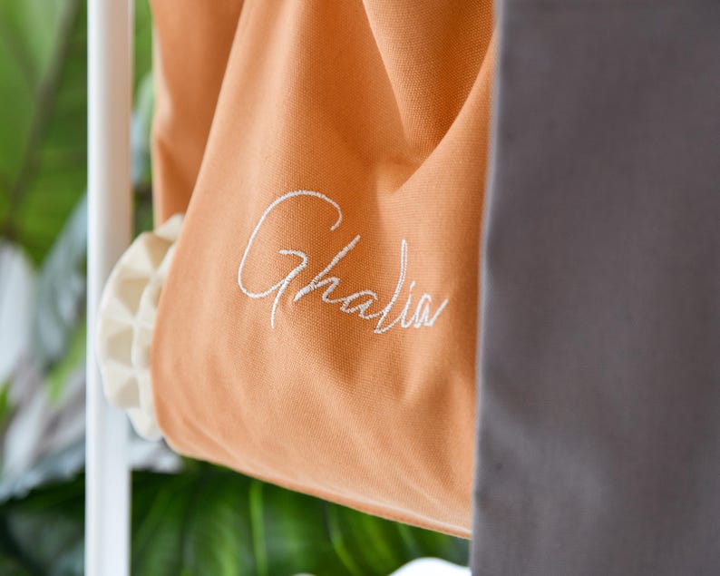 May include: Close-up of an orange canvas bag with the name "Ghalia" embroidered in white script. The bag has a ruffled, off-white trim and is partially visible, with a gray fabric in the background. The bag is hanging on a white rack.