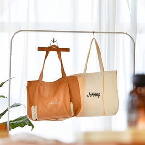 May include: Two tote bags hanging on a white metal rack. One is tan with the name "Ghalia" in cursive, and the other is off-white with "Johnny" in cursive. The bags have long handles and are made of canvas.