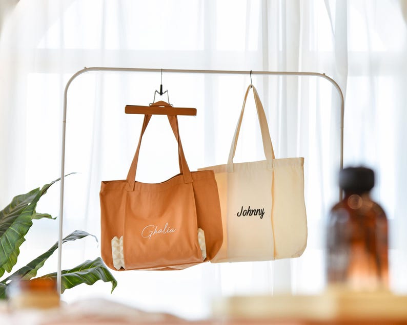 May include: Two tote bags hanging on a white metal rack. One is tan with the name "Ghalia" in cursive, and the other is off-white with "Johnny" in cursive. The bags have long handles and are made of canvas.