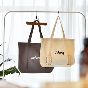 May include: Two tote bags hanging on a metal rack. One is a dark gray color, and the other is cream-colored. Both bags have the name "Johnny" embroidered on them in a cursive font. The cream-colored bag contains two white massage balls.