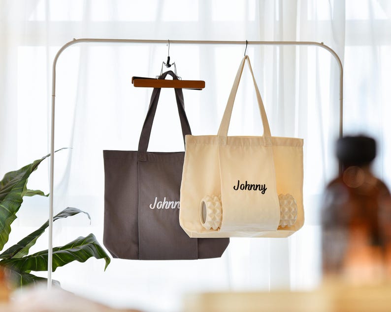May include: Two tote bags hanging on a metal rack. One is a dark gray color, and the other is cream-colored. Both bags have the name "Johnny" embroidered on them in a cursive font. The cream-colored bag contains two white massage balls.