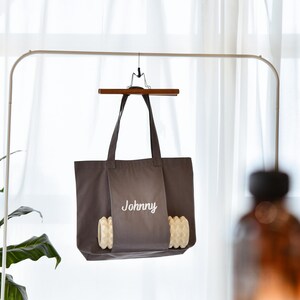 May include: A dark gray tote bag with the name "Johnny" embroidered in white. The bag is hanging from a wooden hanger and has a pocket with a decorative, textured design.