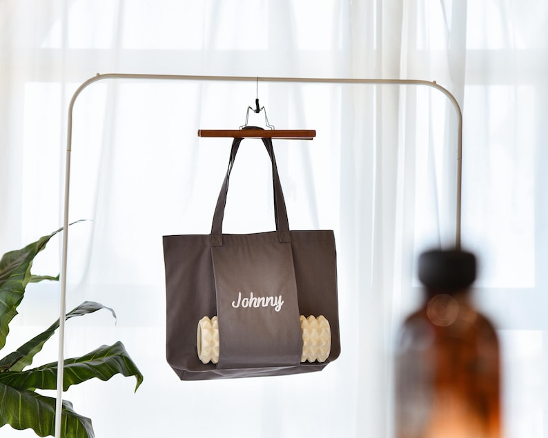 May include: A dark gray tote bag with the name "Johnny" embroidered in white. The bag is hanging from a wooden hanger and has a pocket with a decorative, textured design.