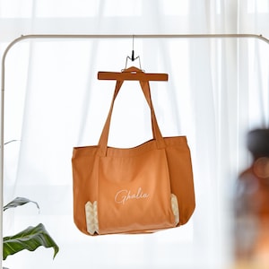 May include: A burnt orange tote bag with long handles, hanging from a wooden hanger. The bag has the name "Ghalia" in cursive script on the front. The bottom corners feature a geometric pattern in white and beige.