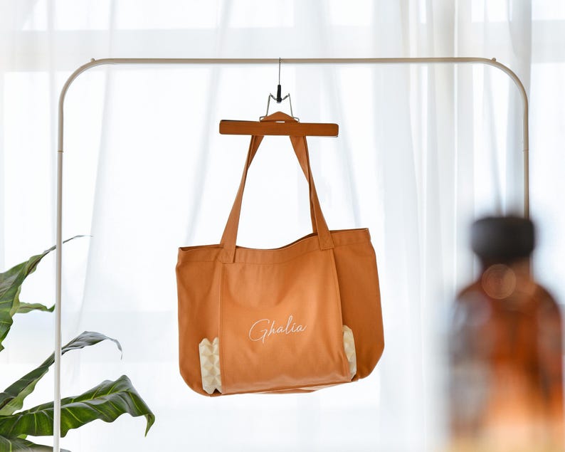 May include: A burnt orange tote bag with long handles, hanging from a wooden hanger. The bag has the name "Ghalia" in cursive script on the front. The bottom corners feature a geometric pattern in white and beige.