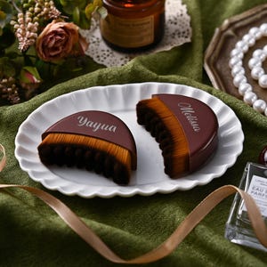 May include: Two brown and tan makeup brushes with the names "Yayua" and "Melissa" printed on them, arranged on a white scalloped plate. The brushes are on a green cloth with a ribbon, flowers, and a perfume bottle in the background.