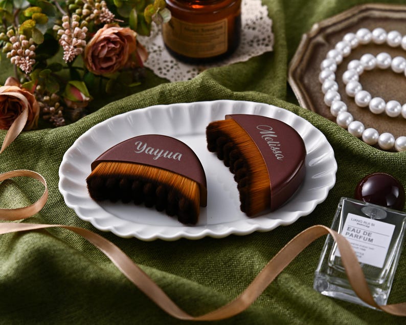 May include: Two brown and tan makeup brushes with the names "Yayua" and "Melissa" printed on them, arranged on a white scalloped plate. The brushes are on a green cloth with a ribbon, flowers, and a perfume bottle in the background.