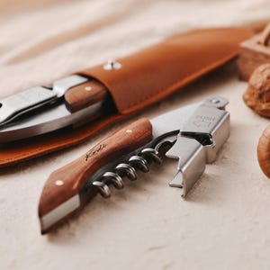 May include: A wooden and stainless steel wine opener with a leather carrying case. The opener has a wooden handle and a silver metal corkscrew. The case is brown leather with a flap.
