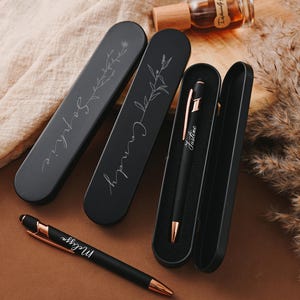 May include: Three matte black elongated pen cases and two matching pens with rose gold accents. Two cases are closed and engraved with floral designs and the names Sophie and Candy. One case is open, containing a pen engraved with the name Justine.