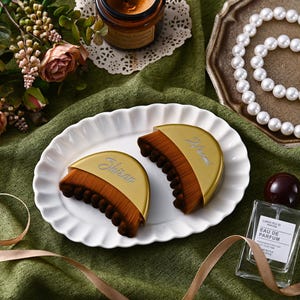 May include: Two gold and brown makeup brushes with the word "Shiraru" and "Narent" printed on them, displayed on a white, scalloped plate. A perfume bottle and a pearl necklace are also visible.