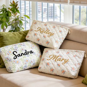 Puede incluir: Tres neceseres acolchados con estampados florales y nombres personalizados: "Kelley", "Sandra" y "Mary". Las bolsas son de color crema con un patrón de diamantes y cierre de cremallera. Los nombres están bordados en oro y negro.