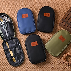 Peut inclure: Une trousse de voyage verte ouverte contenant des couverts, des ciseaux et des couteaux. Plusieurs étuis fermés bleus, noirs et vert olive sont également visibles. Chaque étui a un patch marron. Une caisse en bois et un anneau décoratif en corde sont également dans l'image.