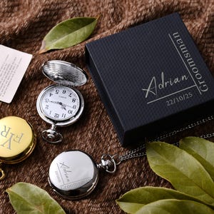 May include: A collection of pocket watches, one open to reveal the clock face, and two closed. One watch is gold-toned with the initials "FYR", and the other is silver-toned with the name "Adrian" and the word "Groomsman". A black box with the name "Adrian" and the word "Groomsman" is also present.