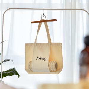 May include: A cream-colored tote bag with the name "Johnny" printed in black script. The bag is hanging on a wooden hanger. Inside the bag are two white and gray patterned items. The background includes a white curtain and a green plant.