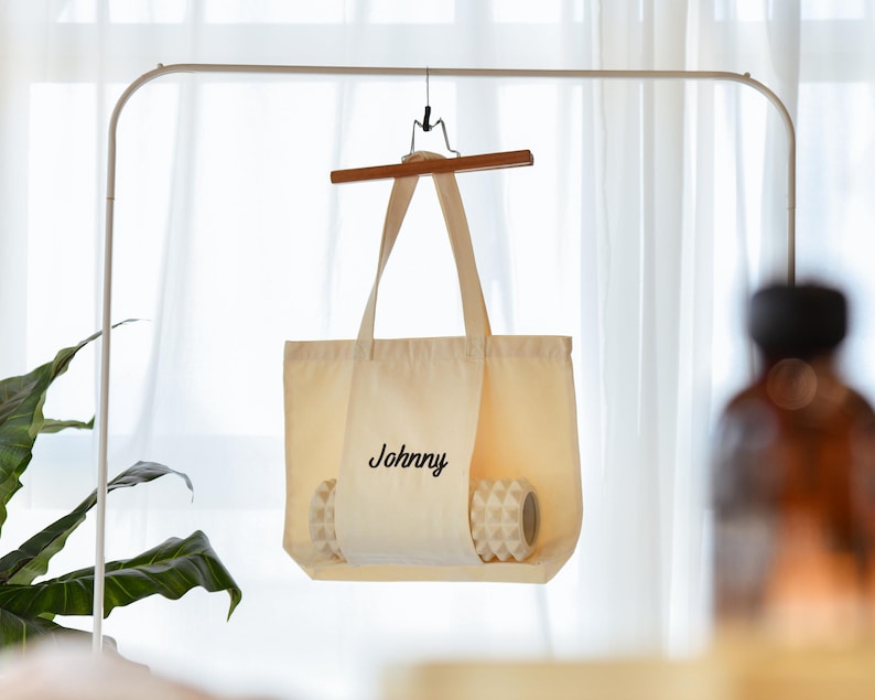 May include: A cream-colored tote bag with the name "Johnny" printed in black script. The bag is hanging on a wooden hanger. Inside the bag are two white and gray patterned items. The background includes a white curtain and a green plant.