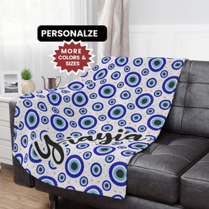 May include: A white blanket with a repeating pattern of blue and black eye-shaped designs. The blanket is draped over a black leather couch. The name "Mayia" is printed on the blanket in a cursive font. The text "PERSONALIZE" and "MORE COLORS & SIZES" are also visible.