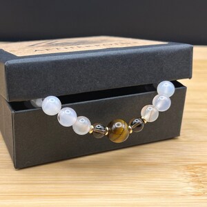 May include: A white agate and tiger's eye beaded bracelet in a black gift box. The bracelet has gold spacers between the beads.