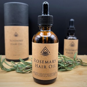 May include: Two amber glass bottles with droppers filled with rosemary hair oil. The bottles have brown paper labels with a logo and the text "Rosemary Hair Oil". The bottles are sitting on a wooden surface with a sprig of rosemary.