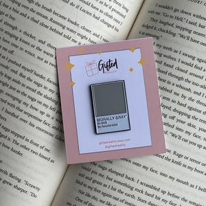 May include: A pin with a gray square and the text "MORALLY GRAY" on a pink card. The card also includes the text "My favorite color" and the Etsy shop's URL. The pin is set against an open book.