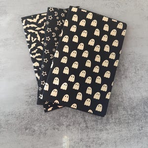 May include: Three rectangular notebooks with black covers featuring repeating patterns in a light cream color. The first notebook displays a pattern of flying bats, the second shows stars and crescent moons, and the third features small ghosts.