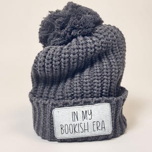 May include: A dark gray knit beanie with a matching pom-pom on top. The hat features a rectangular patch with the text "IN MY BOOKISH ERA" in black lettering. The beanie is made of a thick, textured yarn.