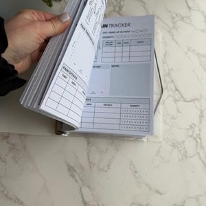 May include: A white and gray pain tracker journal with the text "DAILY PAIN" and "PAIN TRACKER". The journal is open, revealing pages with charts for tracking pain details, severity, and notes. The journal is held open by a hand.