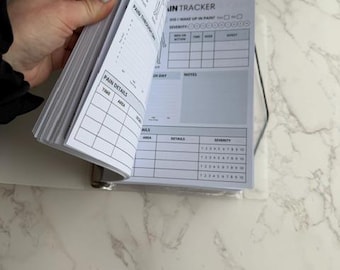 May include: A white and gray pain tracker journal with the text "DAILY PAIN" and "PAIN TRACKER". The journal is open, revealing pages with charts for tracking pain details, severity, and notes. The journal is held open by a hand.